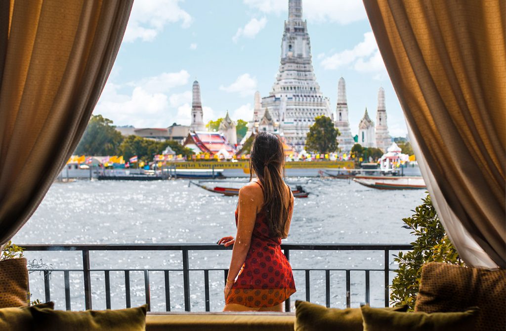 Girl Around the World on the balcony of a hotel room facing a river overlooking Wat Arun