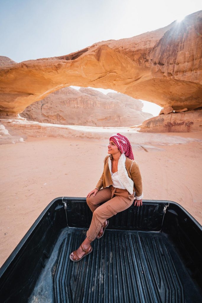 Melissa sits on the back of a Jeep in the Wadi Rum desert. There is a small rock bridge formation behind her.